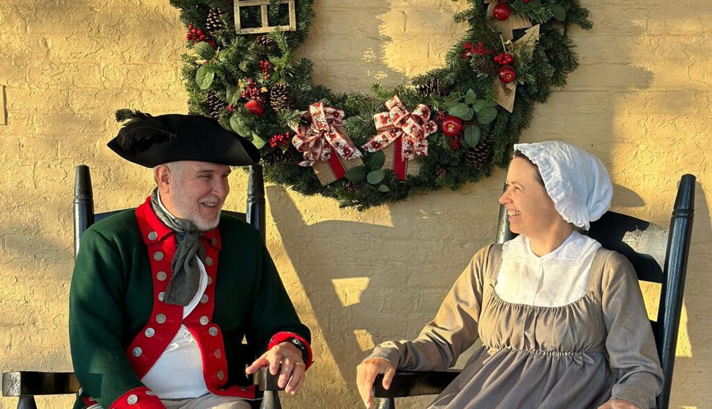 A man and a woman sitting in rocking chairs on a porch in front of a brick wall. They are wearing period (18th century) clothing and the wall behind them features a large christmas wreath. 