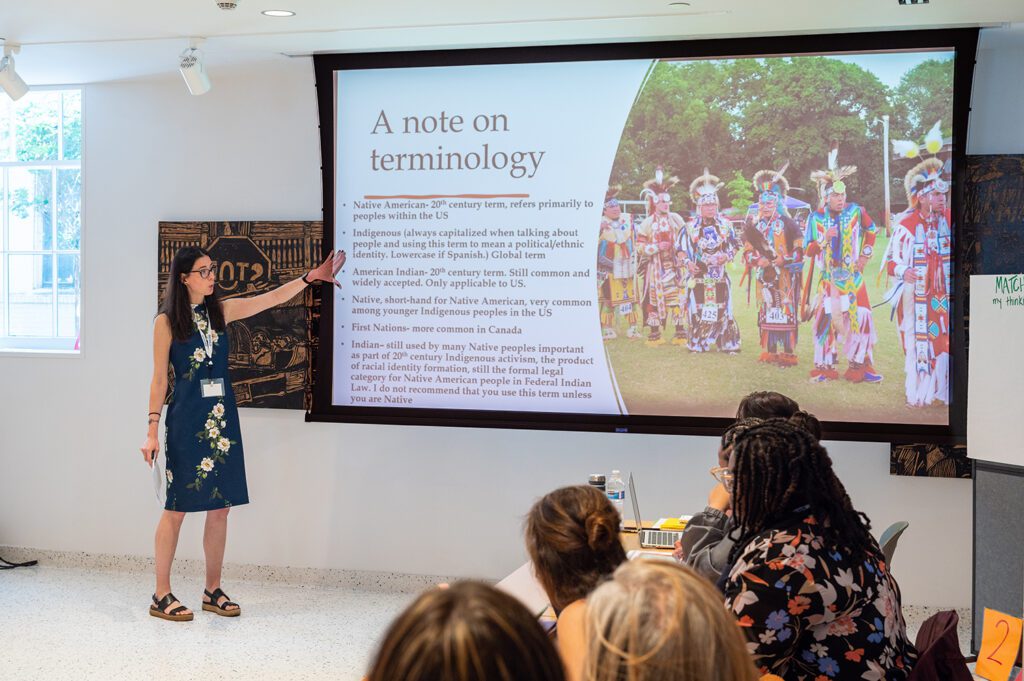 A woman who is a professor stands in front of a large projector screen pointing to the powerpoint presentation which has an image of Native American people in ceremonial dress. Teachers are seated at a table in the foreground of the image watching the professor speak.